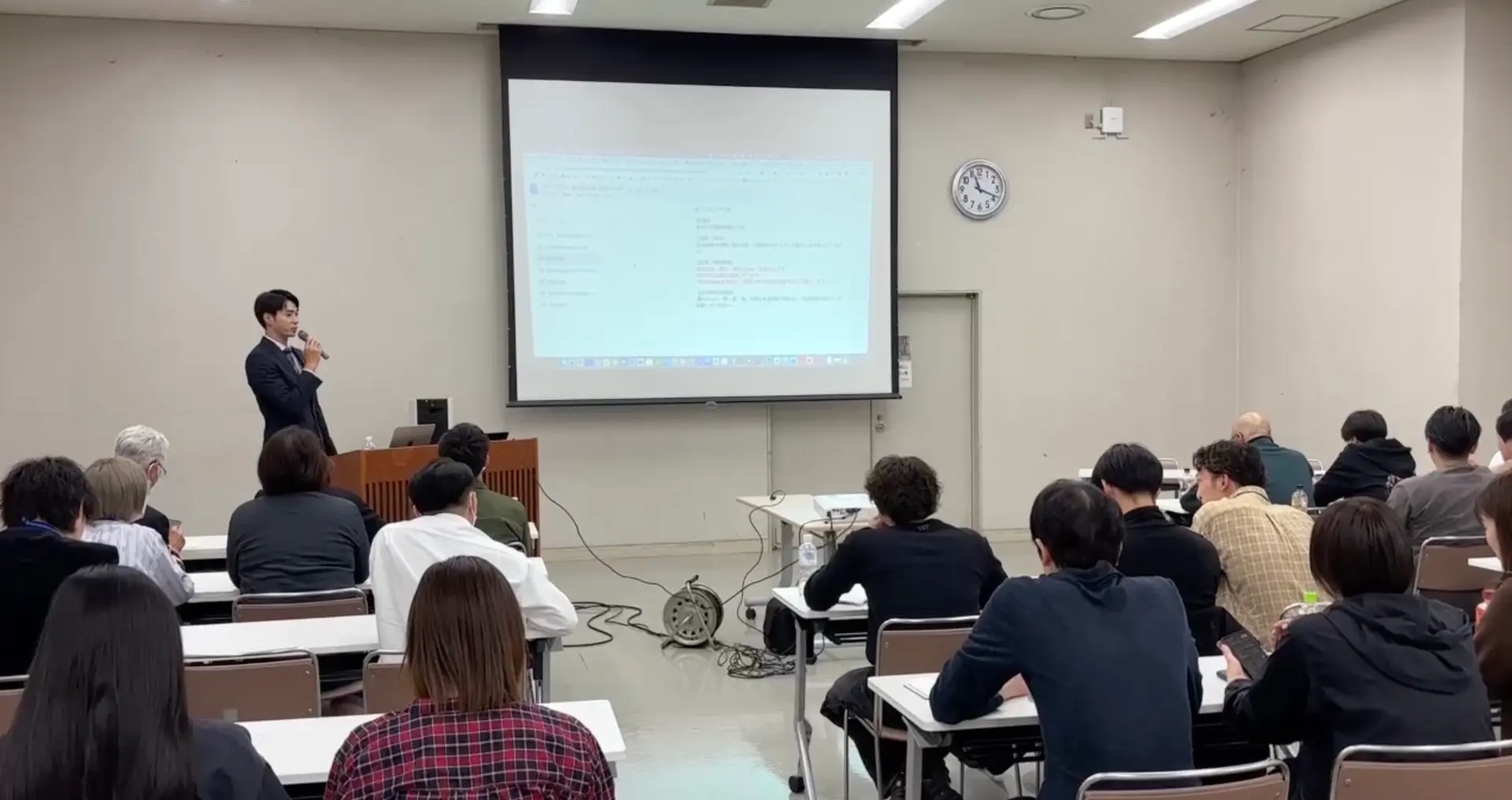 セミナー会場で大勢の参加者に向けて登壇し、スクリーンを使って講演する様子